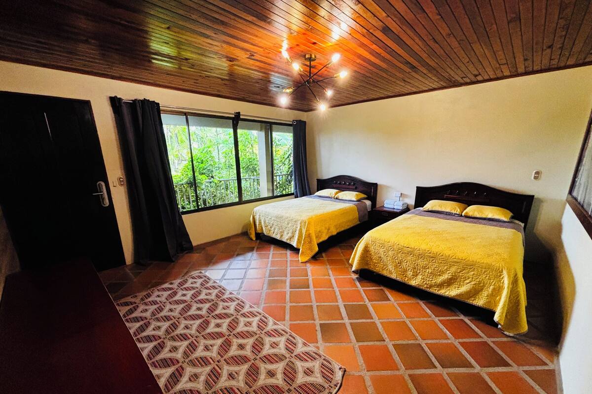 A room with two beds, a wooden ceiling, and a window with a view of greenery.