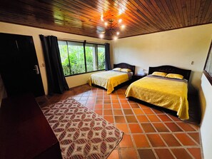 A room with two beds, a wooden ceiling, a window with a view of greenery, and a patterned rug.