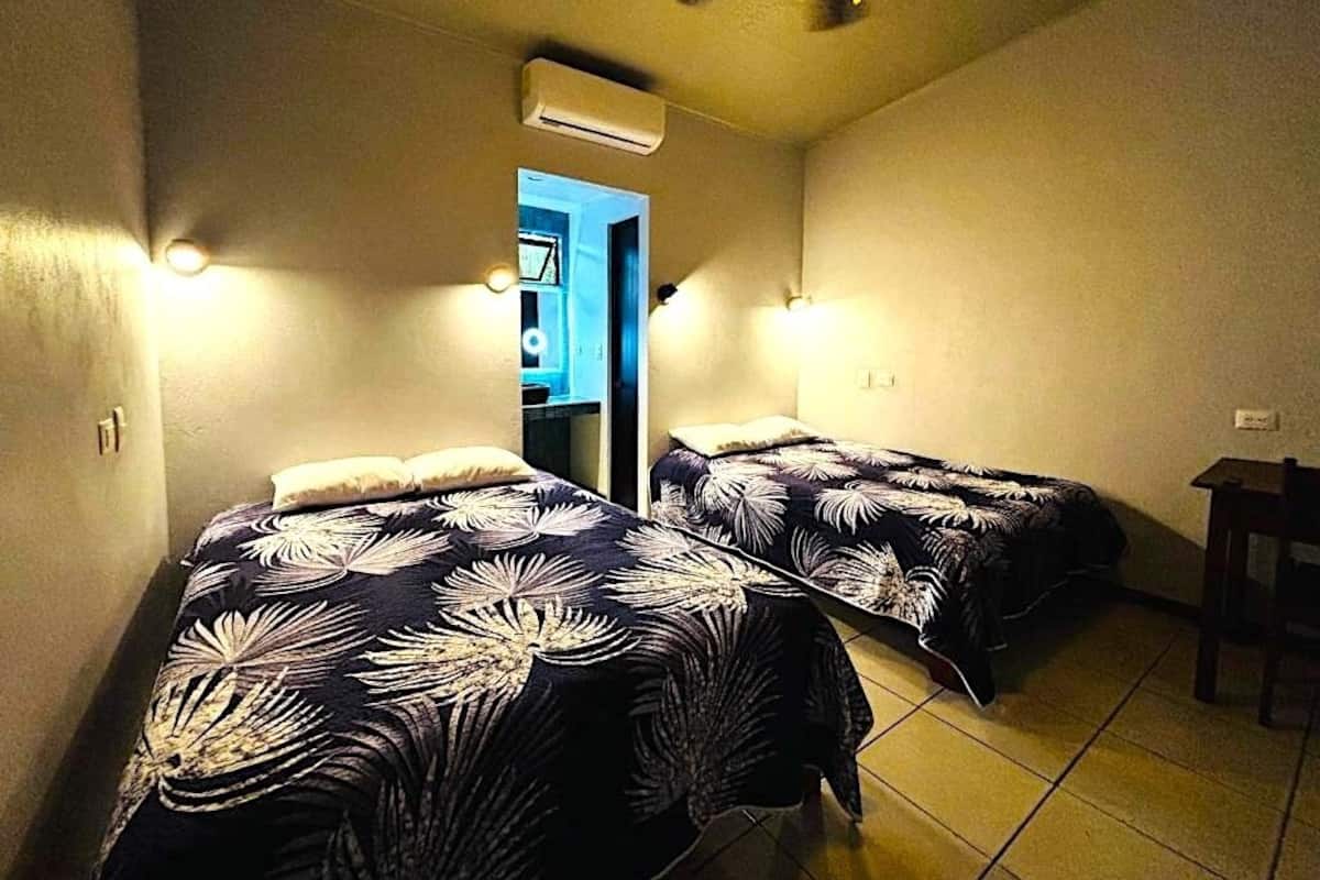 A room with two beds, a ceiling fan, an air conditioning unit, and a door leading to another room.