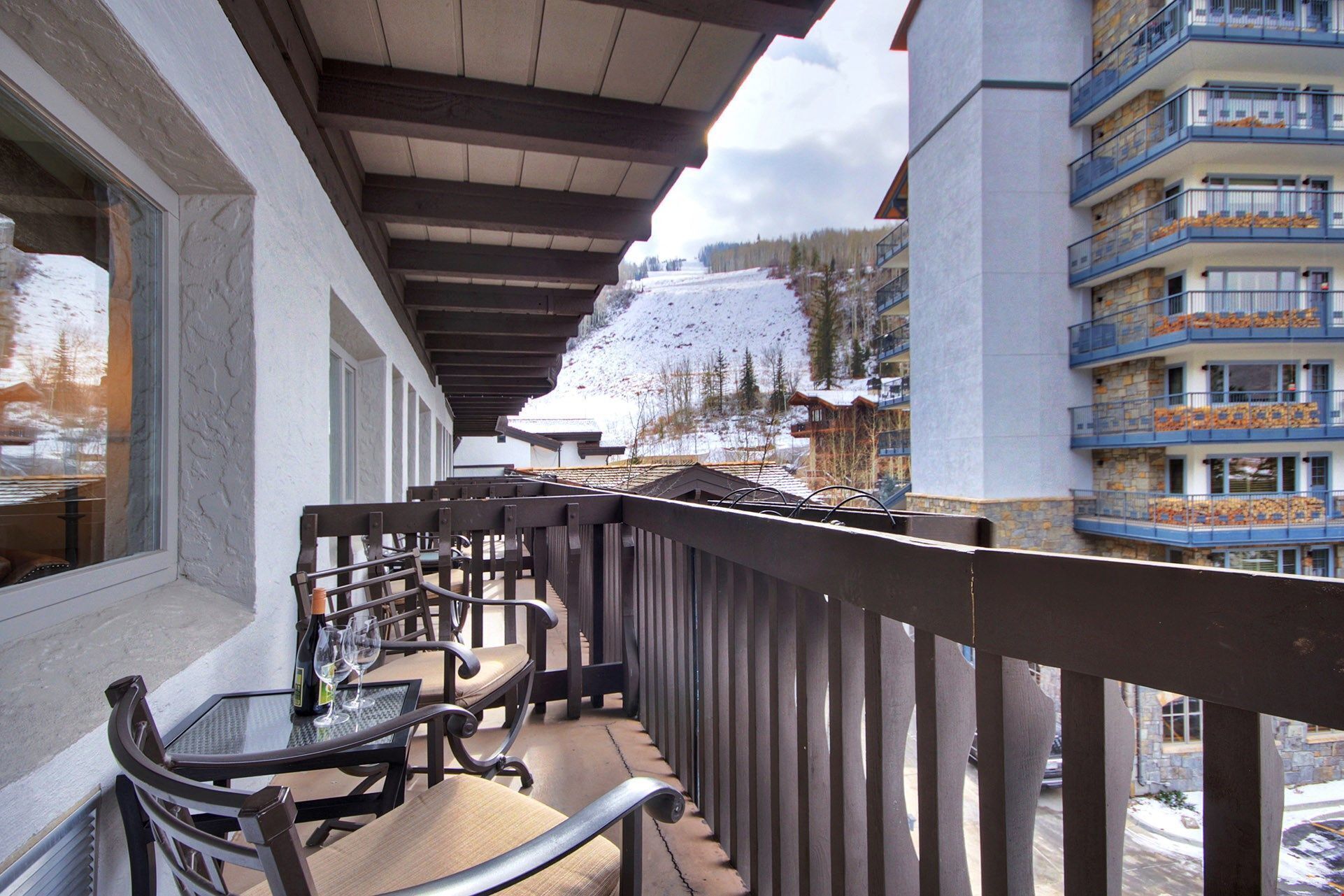 A balcony with a table and chairs, overlooking a snowy mountain and a building with balconies.