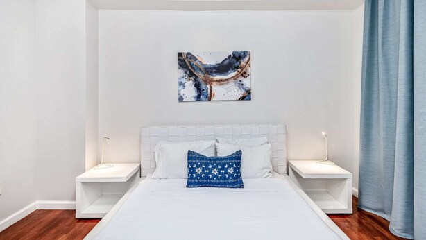 Ein modernes Schlafzimmer mit einem weißen Bett, zwei Nachttischen, einem blauen abstrakten Gemälde und einem blauen Vorhang.