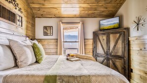 Ein gemütliches Schlafzimmer mit Holzbalkendecke, einem großen Bett mit gemusterten Betttextilien, einem Fenster mit Blick auf die Berge und einem rustikalen Schrank.