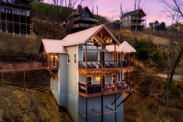 A multi-story house with a balcony and a deck, surrounded by trees and hills.