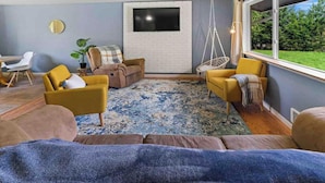 A living room with a TV mounted on a white brick wall, a hanging chair, and a large window with a view of greenery.