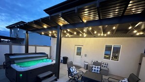 Un bain à remous installé sous une terrasse couverte, agrémentée de guirlandes lumineuses, un coin salon avec des chaises et une table, et une vue sur la maison en arrière-plan.