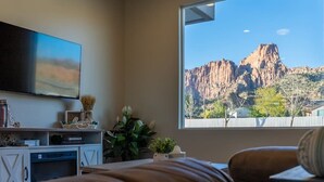Une chambre dotée d’une grande fenêtre offrant une vue sur des montagnes rocheuses, d’un téléviseur à écran plat fixé au mur et d’une table de chevet agrémentée d’objets décoratifs.
