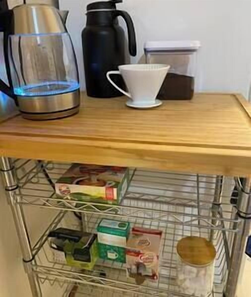 Una encimera de cocina con una cafetera, un hervidor, una jarra de café y un filtro de café. Debajo de la encimera, hay un estante metálico con varios alimentos y un tarro.