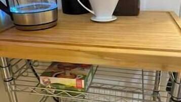 Una encimera de cocina con una cafetera, un hervidor, una jarra de café y un filtro de café. Debajo de la encimera, hay un estante metálico con varios alimentos y un tarro.