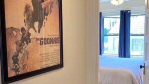 Hotellihuone, jonka seinällä on kehystetty elokuvan juliste, valkoisilla lakanoilla pedattu sänky ja ikkunasta avautuva näkymä ulos, ikkunassa on verhot.