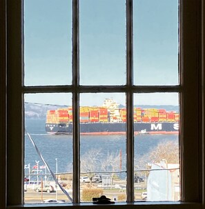 Een groot vrachtschip met meerdere containers in het water, zichtbaar door een raam met een rasterpatroon.