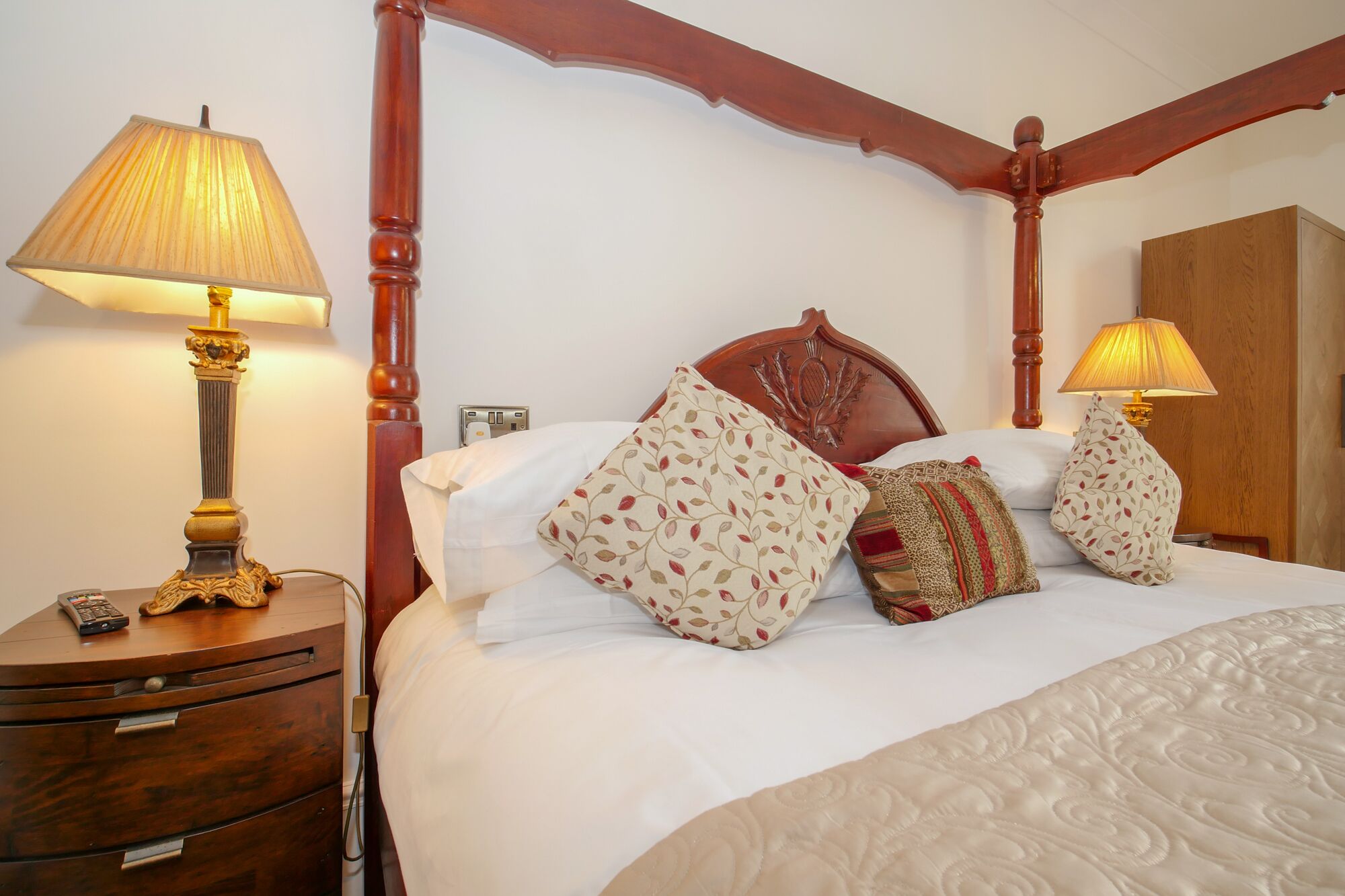 A four-poster bed with a white bedspread and multiple pillows, including floral and patterned designs. There are two bedside tables with lamps, one of which has a remote control on it. The room has wooden trim and a wooden headboard.
