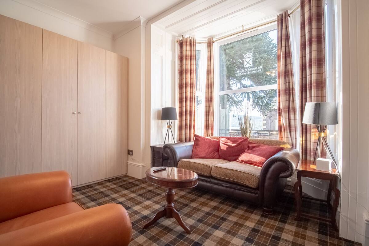 Un salon comprenant un canapé, une table basse, une lampe et une vue sur les arbres à travers une fenêtre ornée de rideaux.