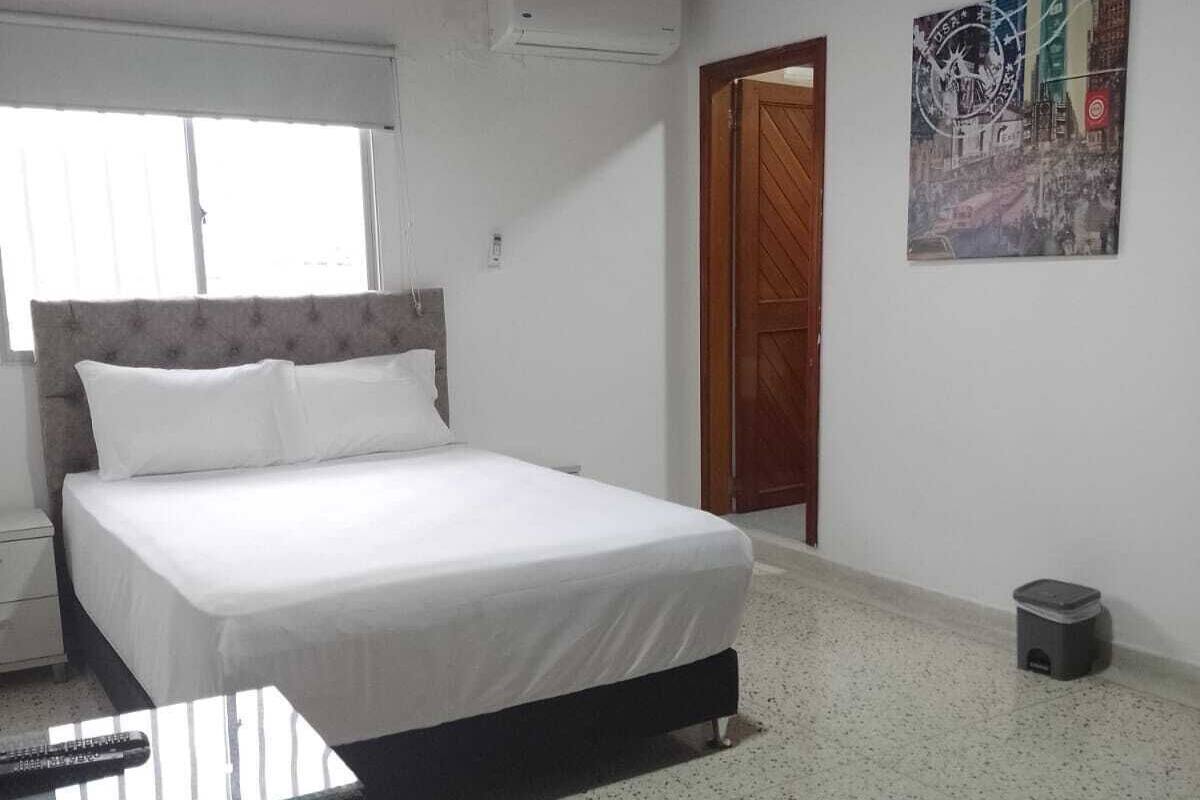 A bedroom with a bed, a small table, a window, an air conditioning unit, a door, and a trash bin.