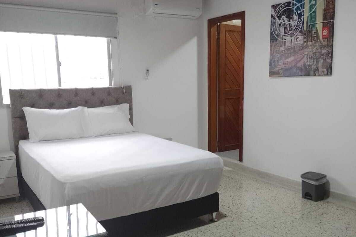 Un dormitorio con una cama, una mesita, una ventana, un equipo de aire acondicionado, una puerta y un cesto de basura.