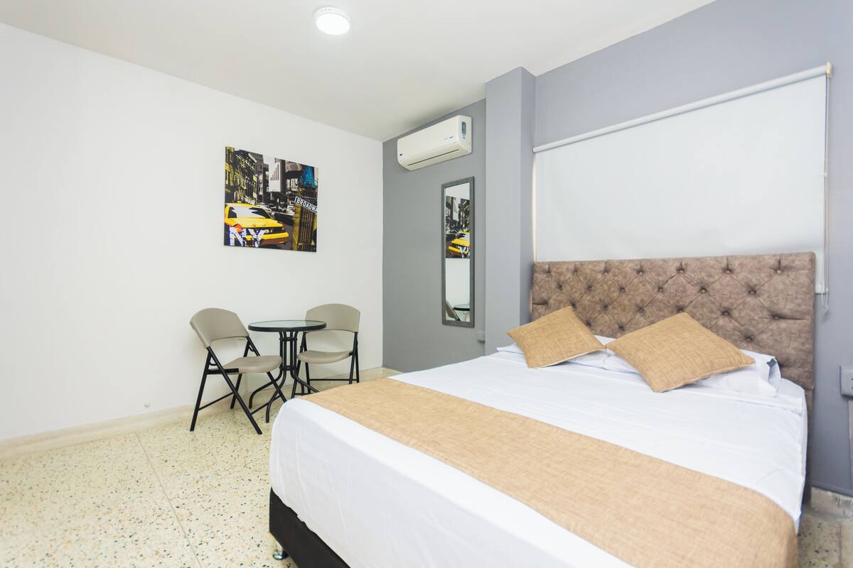 A hotel room with a large bed, a small table, two chairs, and a wall-mounted air conditioner.