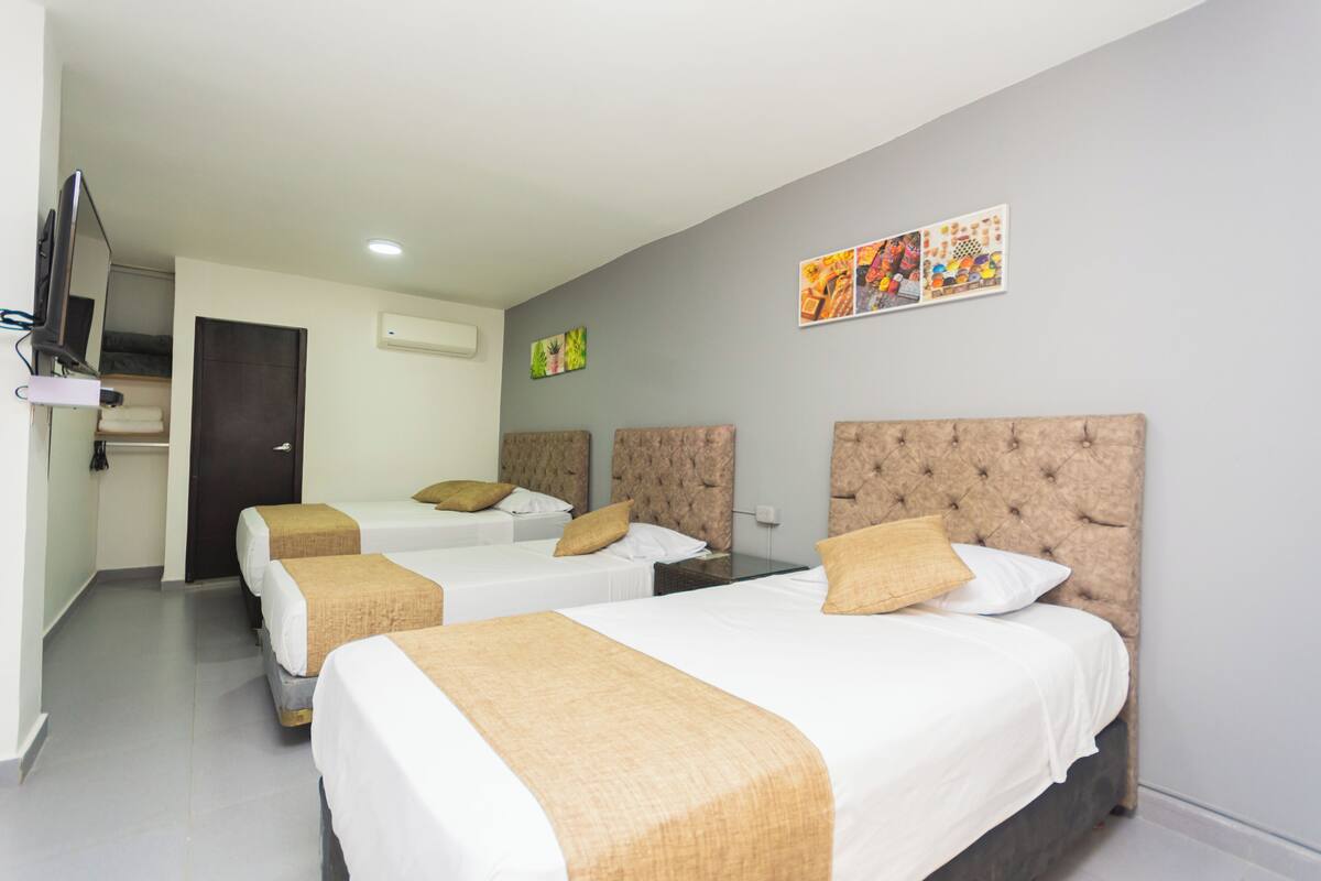A hotel room with three beds, each with a headboard and a beige blanket. There is a flat-screen TV mounted on the wall, and artwork is displayed above the beds.