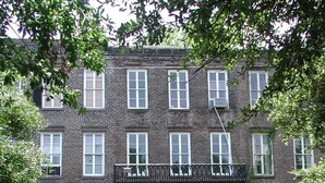 A multi-story brick building with white-framed windows, a black metal balcony, and a car parked in front.
