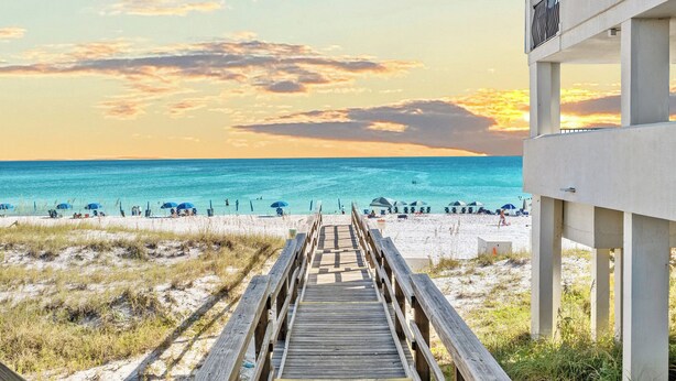 A wooden boardwalk leading to a beach with umbrellas and people.