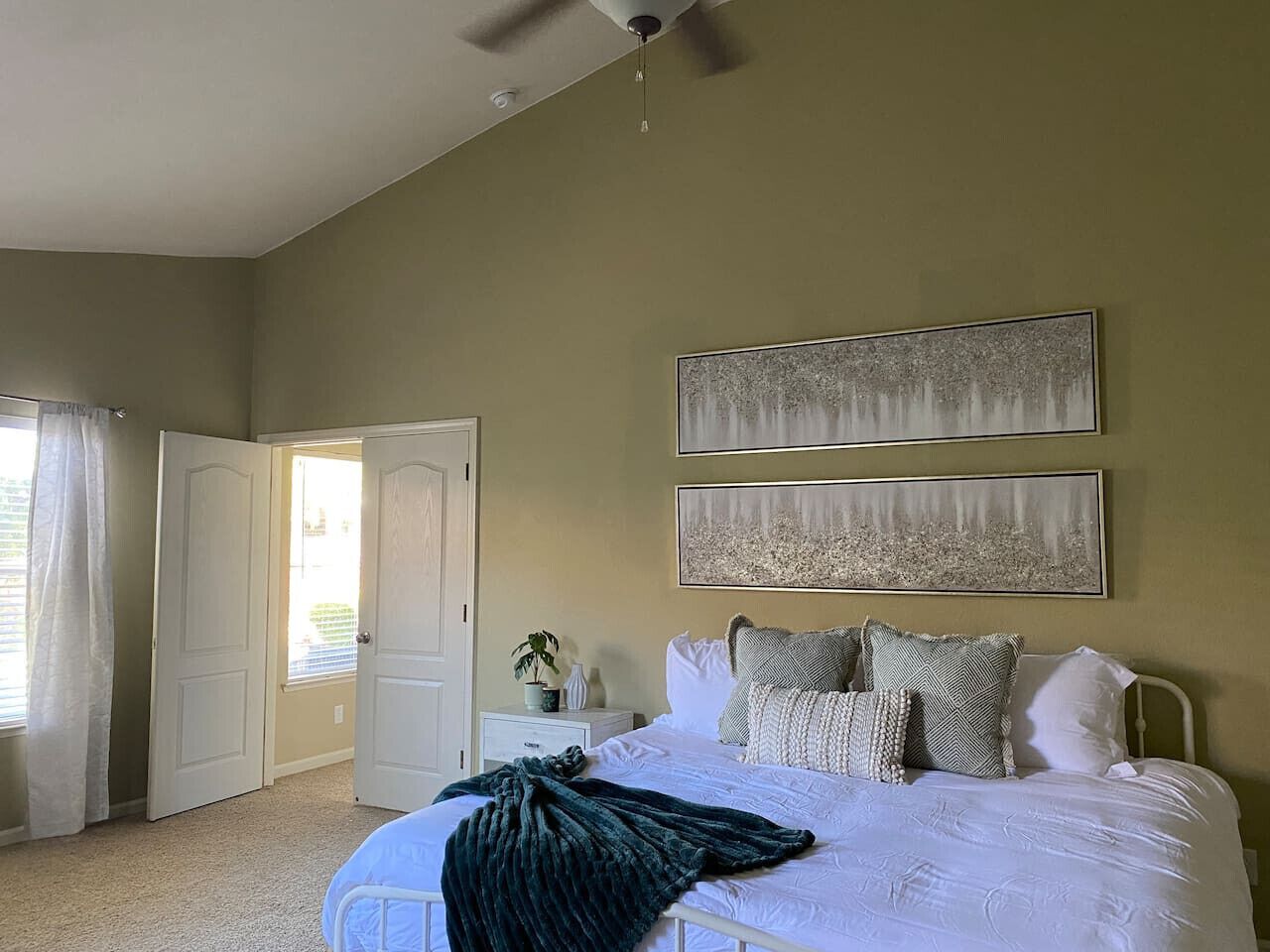 A bedroom with a bed, two paintings, a nightstand, a potted plant, and a door to another room.
