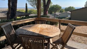 A wooden deck with a round table and four chairs, overlooking a scenic landscape with trees and a view of the sea. - Chalet pour 6 proche Commerces et Plage (Agde)