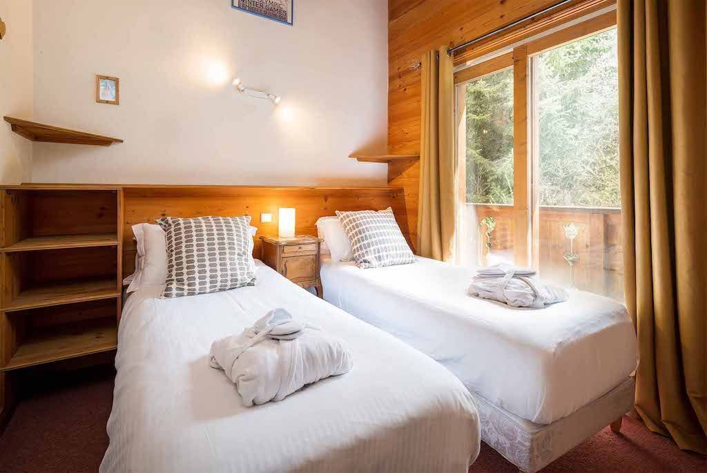 Una habitación con dos camas individuales, cabeceros de madera, una estantería y un ventanal con cortinas.