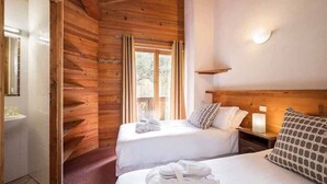A wooden cabin-style room with two beds, a window with curtains, a bedside table with a lamp, and a wall-mounted shelf.