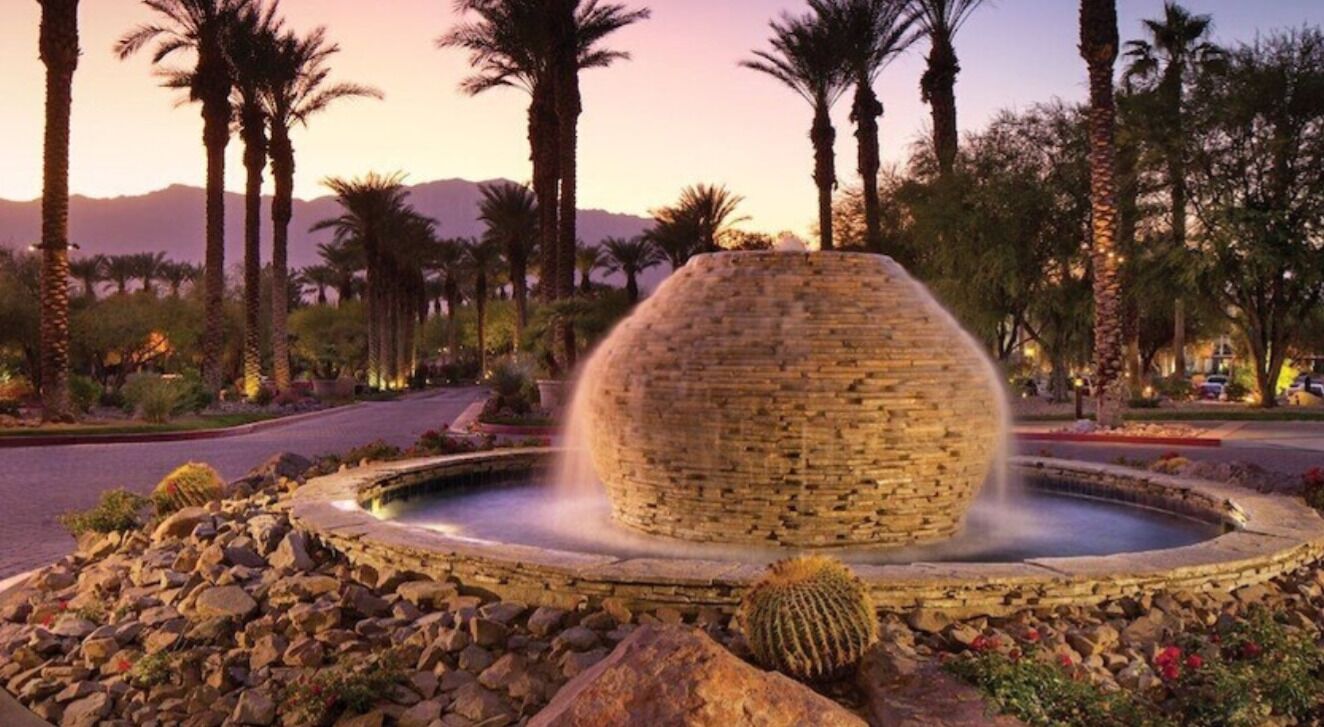 Een ronde stenen fontein met waterstralen, omringd door rotsen en cactussen, gelegen in een aangelegd gebied met palmbomen en een berg op de achtergrond.