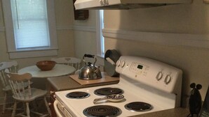 Een keuken met een witte kookplaat, een waterkoker en een kom op het aanrecht. Er is een raam met luiken en een stoel.