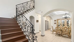 Eine Treppe mit aufwendiger schwarzer Geländer, ein Essbereich mit Holzsesseln und einer Glasvitrine sowie ein Chandelier.