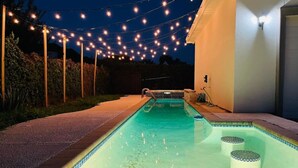 Ein Pool mit Lichterketten darüber, umgeben von einem gepflasterten Weg und einem Gebäude.