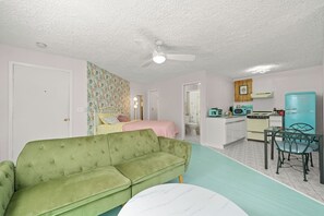 Ein gemütlicher, kompakter Wohnbereich mit einem grünen Sofa, einem runden Tisch, einer Küchenzeile mit einem türkisfarbenen Kühlschrank und einem Deckenventilator.