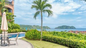 A poolside area with a palm tree, a table with chairs, and a view of the sea and distant islands. - Amazing Ocean Views: Luxury Apartment @ Los Sueños (Playa Herradura)