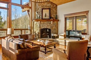 A cozy living room with a stone fireplace, leather sofas, and a large window overlooking a forest. - Gorgeous Summit County Family Oasis (Silverthorne)