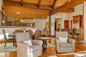 A spacious living room with two armchairs, a dining area with a chandelier, and a kitchen with wooden cabinets.