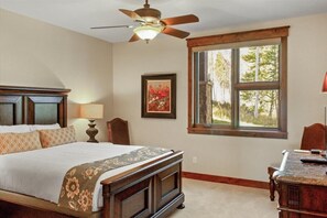 A bedroom with a large bed, a ceiling fan, a window with a view of trees, and a desk with a lamp.