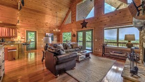 Ein gemütliches Blockhauss-Interieur mit Holzboden, einer Ledersofa, einer Essecke und großen Fenstern mit Blick ins Freie.