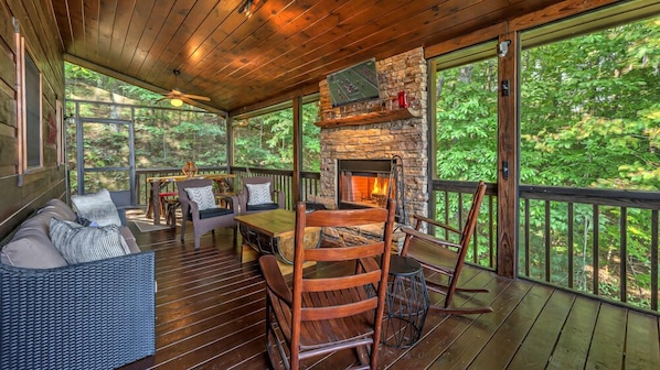 A wooden deck with a stone fireplace, seating area, and large windows overlooking a forest.