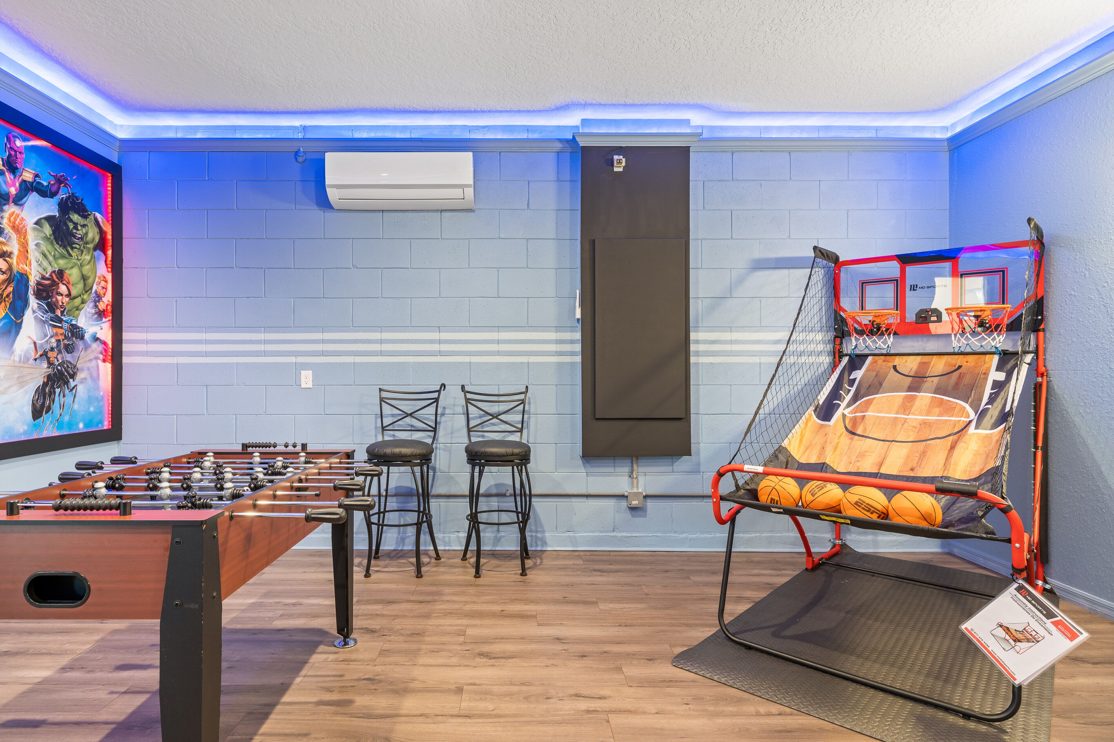 A room with a foosball table, two chairs, a basketball hoop, and a large TV screen displaying a sports game.
