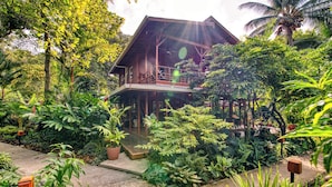 A wooden house with a balcony, surrounded by lush greenery and tropical plants.