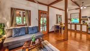 A room with a sofa, a wooden coffee table, a dining area with a table and chairs, and a kitchen with a microwave and sink.