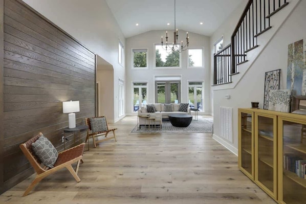 A spacious living room with a high ceiling, a staircase, and a large window allowing natural light.