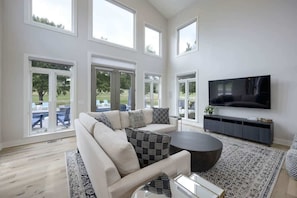 A modern living room with a sofa, coffee table, and TV. Large windows offer views of the outdoors.