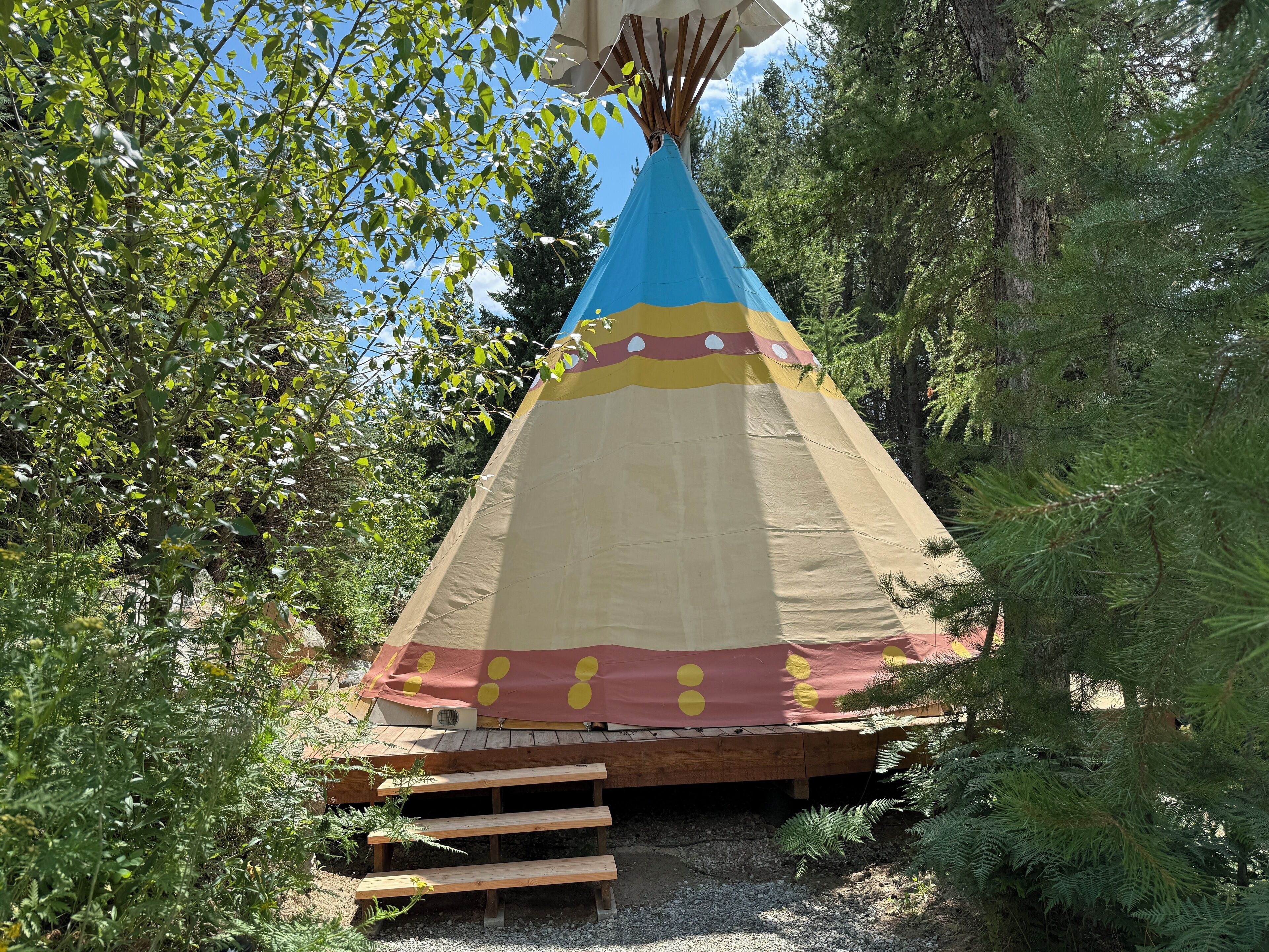 Uma tenda cónica com um design colorido, rodeada por árvores e uma plataforma de madeira.