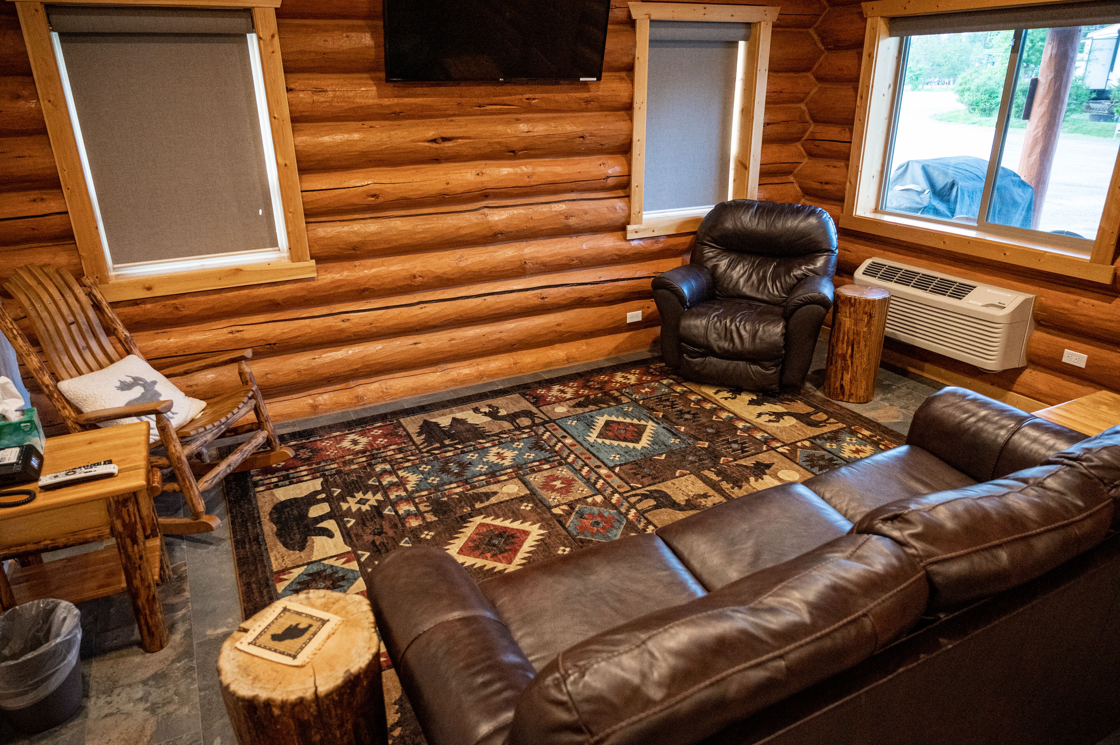 Interior de una cabaña de troncos con un sofá y un sillón de cuero, una mesa de madera y una alfombra estampada.