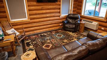 Interior de una cabaña de troncos con un sofá y un sillón de cuero, una mesa de madera y una alfombra estampada.