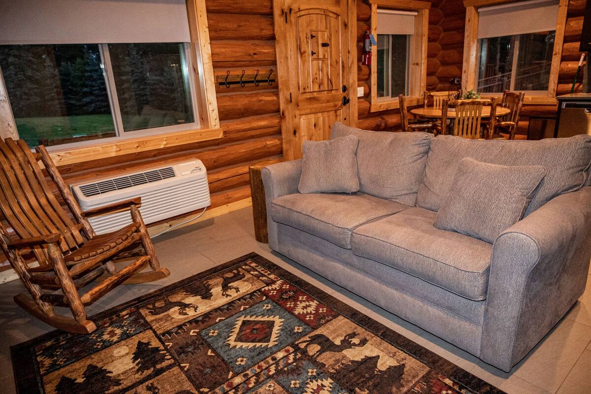 A log cabin interior with a gray sofa, wooden rocking chair, and a patterned rug.