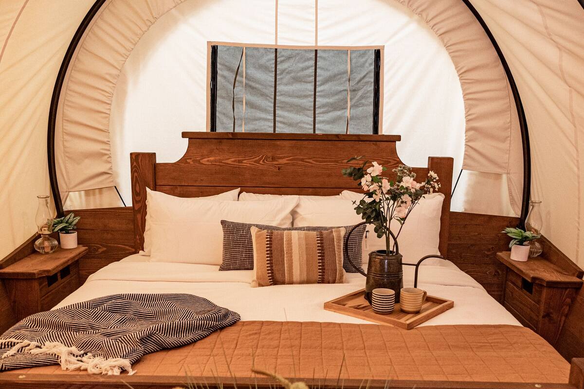 A tented room with a wooden bed, a bedside table, a hanging light, and a window.