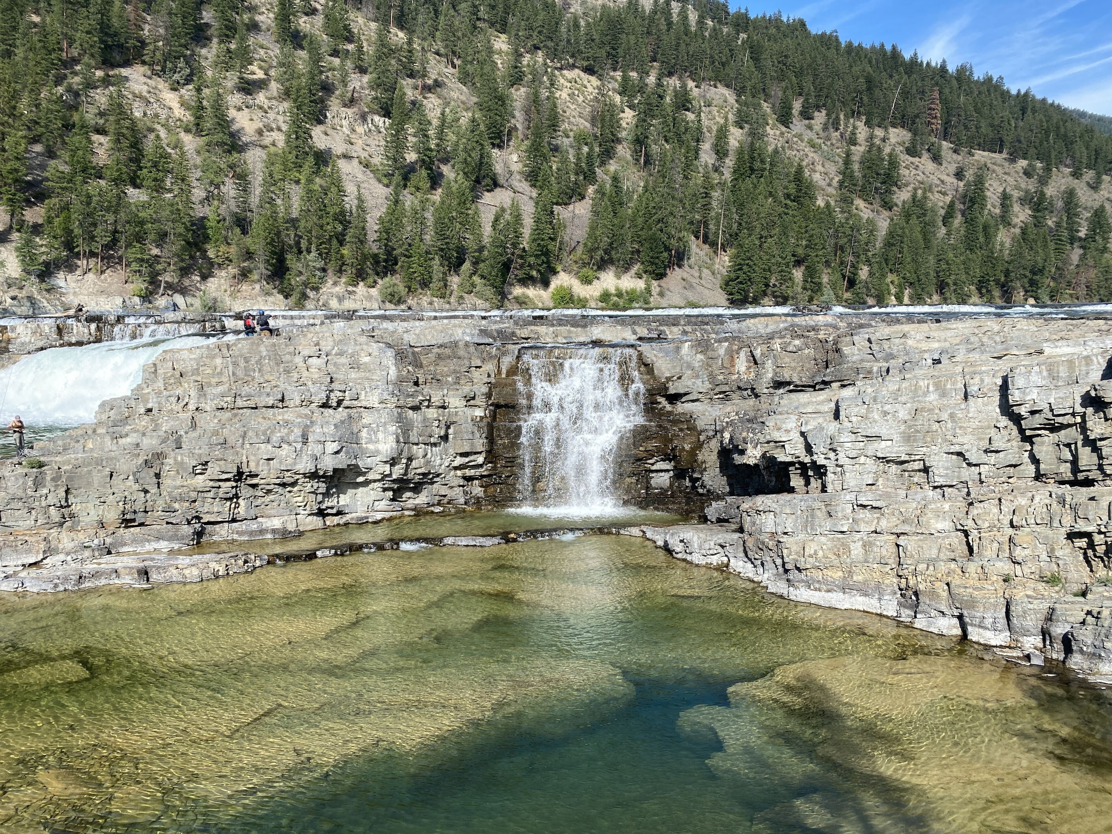 A natural landscape with a waterfall, clear water, and a forested hillside.