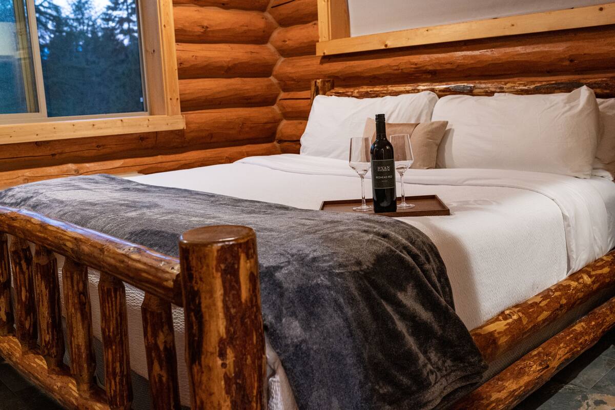 A wooden log cabin room with a bed, a bottle of wine, and two glasses on the bed.