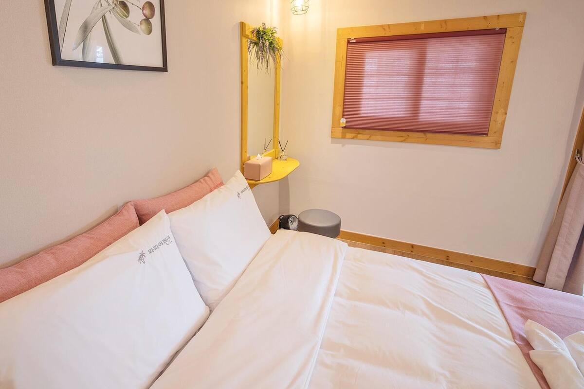 A neatly made bed with white and pink bedding, a small bedside table with a plant, a mirror, and a window with a wooden frame.
